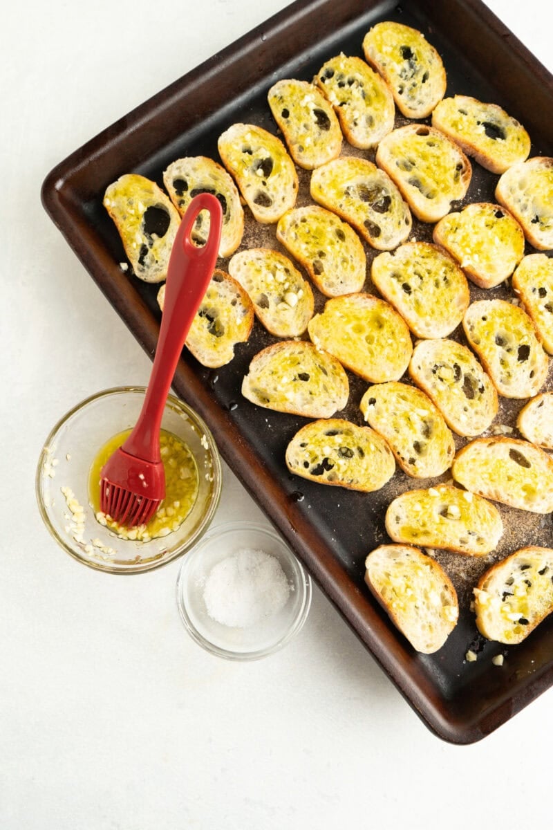 Baguette slices on a baking sheet being basted with garlic and olive oil and sprinkle with salt. Then bake.