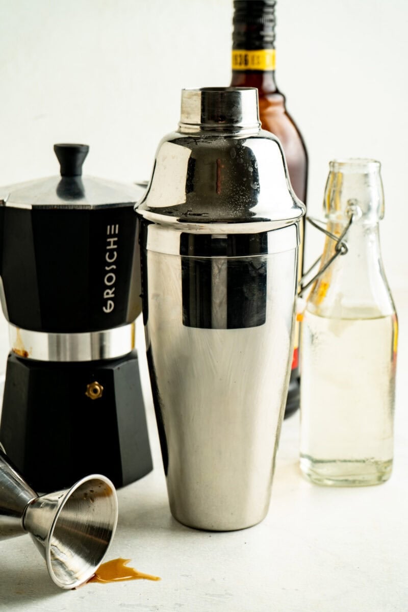 A cocktail shaker on a table with simple syrup and an espresso pot.
